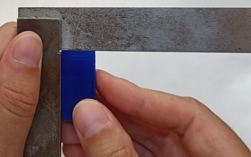 Close up of hands holding a set square and measuring a blue piece of wax in the corner. Both sides are touching 