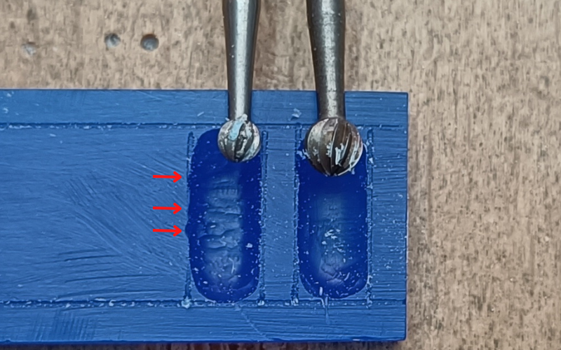 Close up of a piece of blue wax on a bench peg. 2 rectangles are marked on the wax and they're partially burred. In one rectangle lies a ball burr that fits nearly perfect between the lines the burred surface is pretty smoooth, in the other lies a ball burr that's a lot smaller than the lines the surface is not as smooth in this one. The burred indent done by the big burr is straight and follows the marked line. The burred indent doen by the smaller burr has a few outliers, red arrows point top them