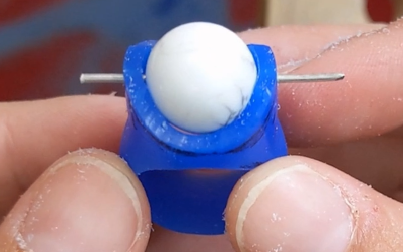  Close up of hands holding an in process blue wax ring. A bead is suspended inside the ring with some metal wire going through ring and bead to hold it in place