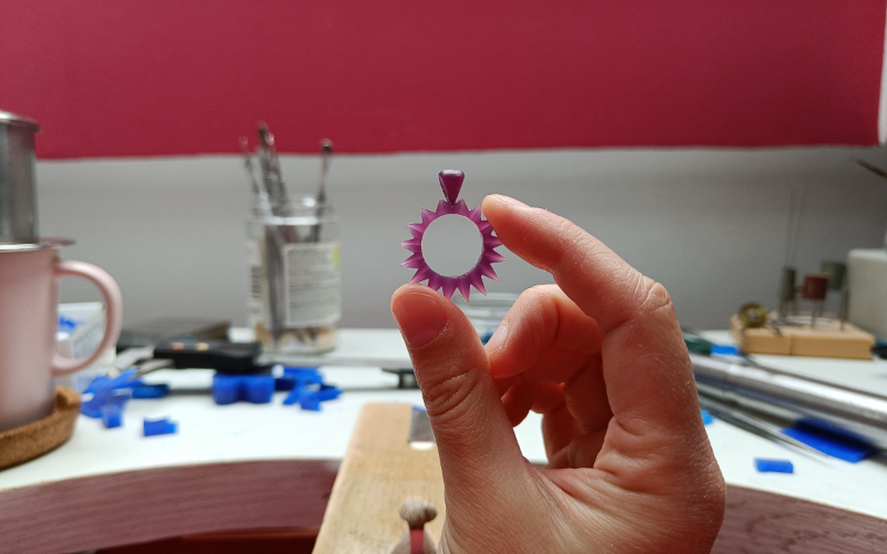 Close up of hands holding a pendant from purple wax. In the background you can see the bench with a lot of distractions on it