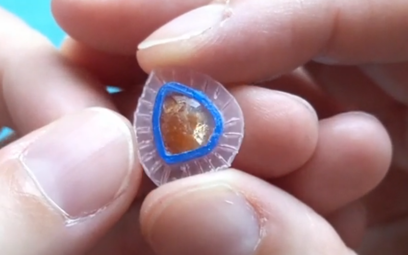 Close up of hands holding a small bezel for a freeform sunstone. The pink sheet backplate is textured with lines radiating from the center outwards