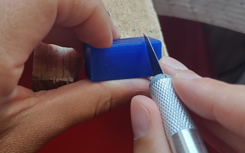 Left hand holding a square piece, right hand holds a stanley knife against the wax. Left hand is below the stanley knife, in danger of being cut  