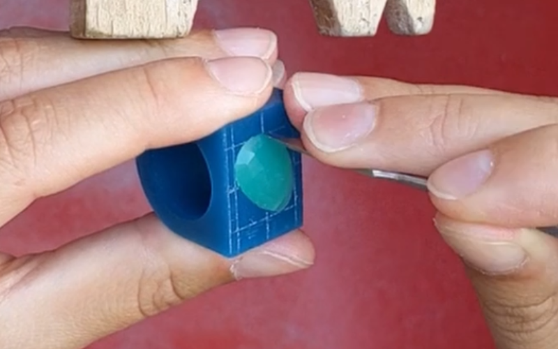 Closeup of hands working at the bench. A turquoise wax ring with a stone inside. The stone is stuck in the setting so a thin scalpel is pushed between stone and setting wall