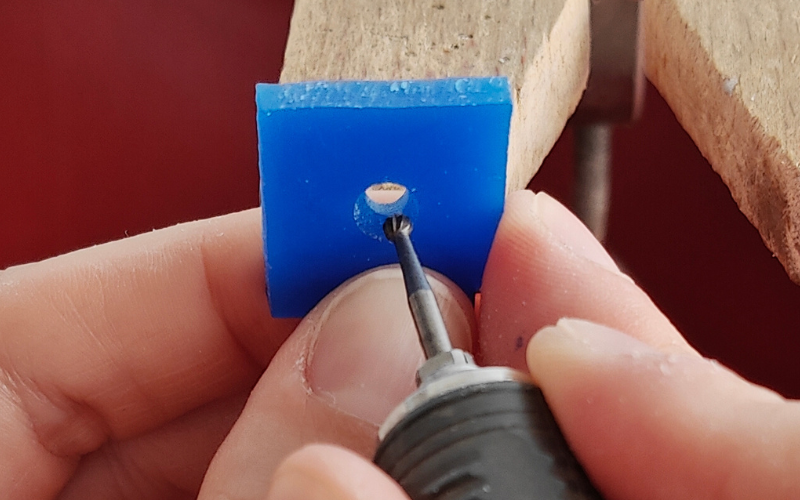 Close up of hands holding a piece of blue square wax with a hole in the center. The piece is angled towards the camera and the other hand is burring the bottom quarter of the hole. You can't see the top quarter and the sides are partially obscured as well 