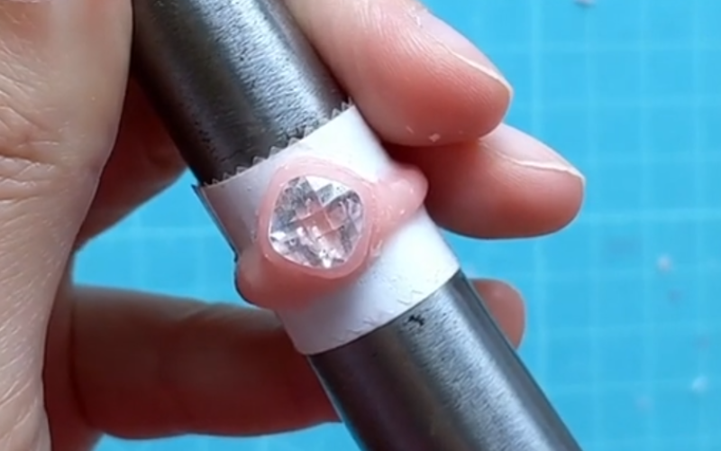 hands holding a ring mandrel with a ring on it made from pink sheet wax and a cushion cut white cz in the center of the ring