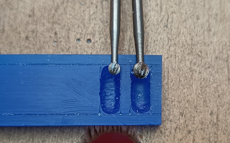 Close up of a piece of blue wax on a bench peg. 2 rectangles are marked on the wax and they're partially burred. In one rectangle lies a ball burr that fits nearly perfect between the lines the burred surface is pretty smoooth, in the other lies a ball burr that's a lot smaller than the lines the surface is not as smooth in this one. 