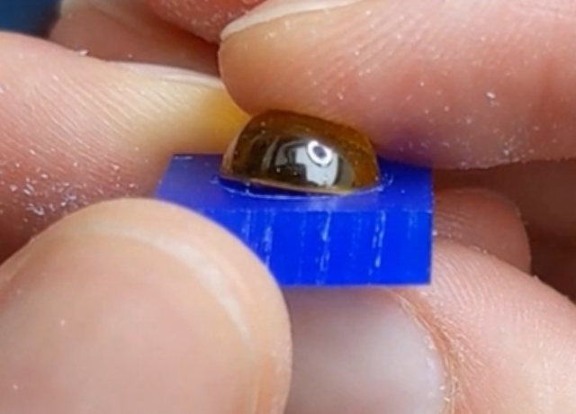 Close up of hands holding a piece of blue wax with a yellow round cabochon on top. The cabochon is partially inside the openiong that's been carved and partially sticking up