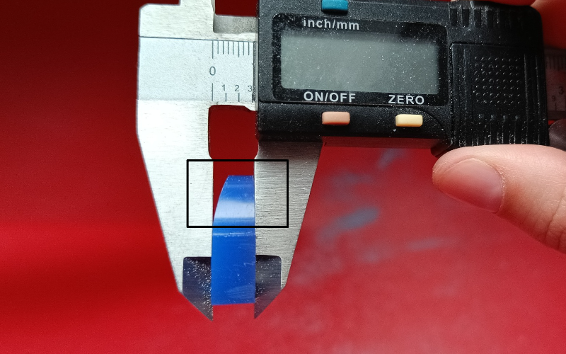 Close up of hands holding a piece of blue ring wax in calipers. A black rectangle is drawn around the place you should look at 