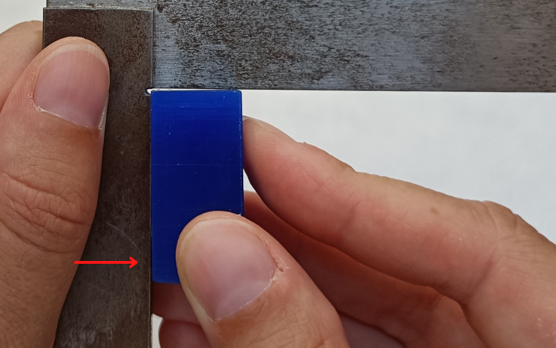 Close up of hands holding a set square and measuring a blue piece of wax in the corner. A red arrow points to the place where I'm angling the wax away from the set square.