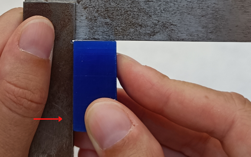 Close up of hands holding a set square and measuring a blue piece of wax in the corner. A red arrow points to the side that is completely touching the arm of the set square