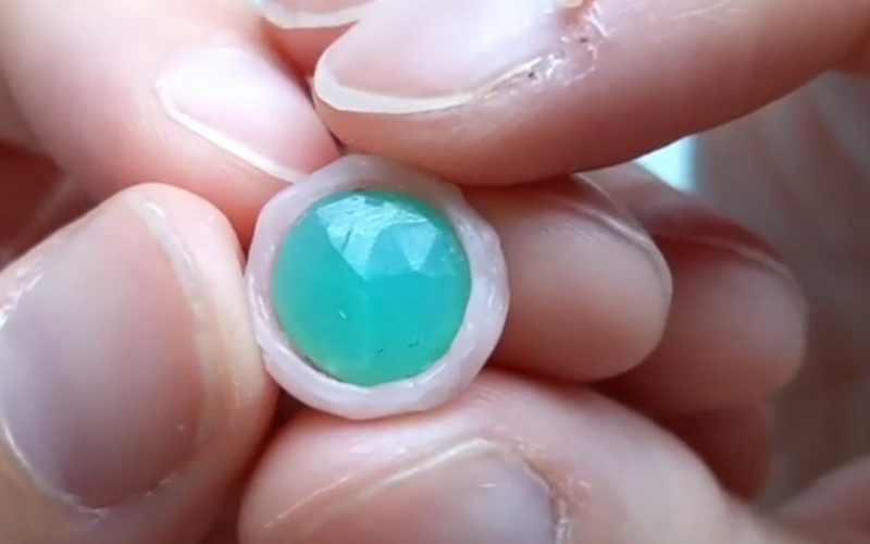 Close up of hands holding a chrysoprase with a finished bezel from pink sheet wax visible around it