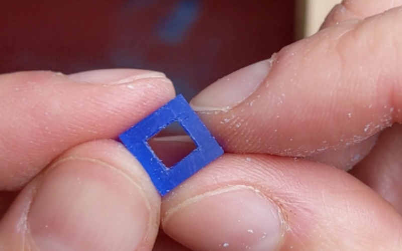 Hands showing a piece fo wax that now has the shape perfectly pierced to a square.