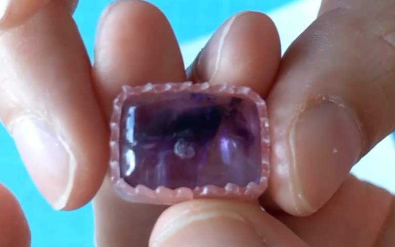 Close up of hands holding a big rectangular amethyst with a pinksheet wax bezel around it. The edges of the bezel are serated