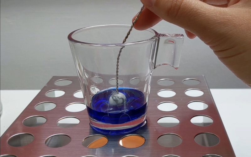  Close up of a small glass on top of a tealight. Blue wax is completely melted in the glass. A hand is holding a bit of wire with a bead at the end, the bead is being dipped in the liquid wax