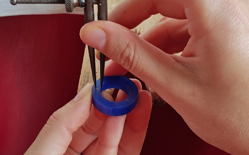Close up of hands holding a piece of blue ring wax. Right hand is marking a line with dividers using the inside of the ring as a guide. Dividers are straight and the point of the guiding leg of the divider is near the top