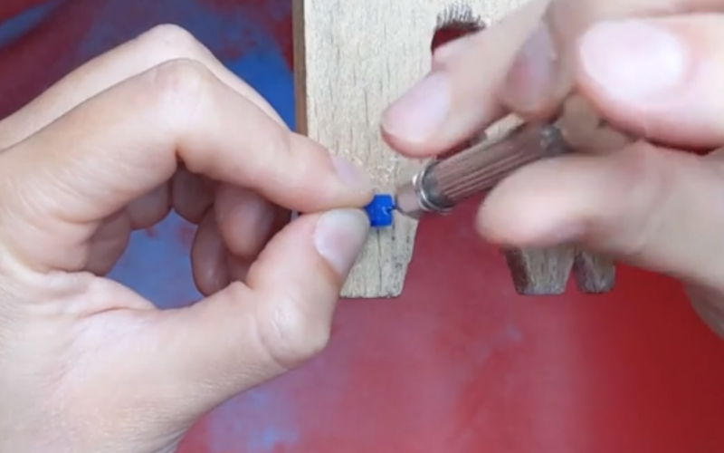 close up of hands working at the bench. A small piece of blue wax lies on the peg and hands are manually drilling through the wax with a drill bit in a pin vice