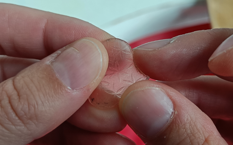 Close up of hands holding a piece of pink sheet wax. Fingernail is scraping around the edge of the wax to smoothen it