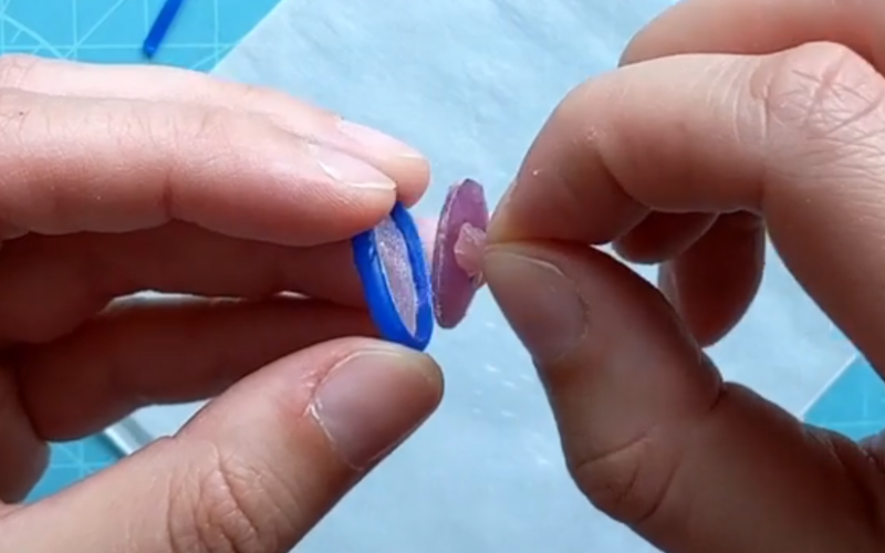 close up of left hand holding a bezel from pink sheet and blue wire wax for an oval stone while the right hand pulls out an oval sapphire stone from the bezel
