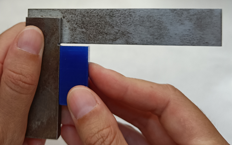 Close up of hands holding a set square and measuring a blue piece of wax against the thick arm. There are no gaps visible