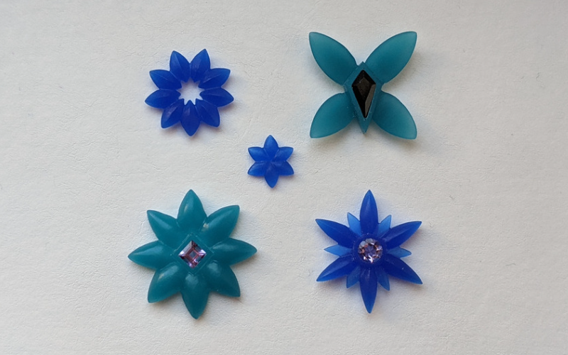Flat lay of 5 different flower shapes in wax. One blue wax flower shape with pointed petals and an open centre, one turquoise wax flower with 4 petals and a black kite shaped stone in the centre. One very small blue wax 6 petal flower. One turquoise wax flower shape with a square purple centre stone, one blue wax flower shape with long and short petals and a round purple centre stone