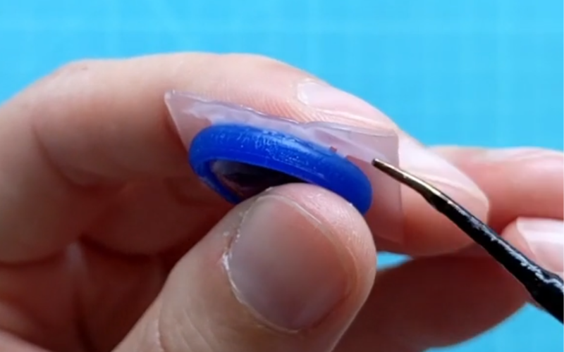 Close up of hands holding a piece of pink sheet wax with a piece of blue wax wire that's half melted to the surface. A small gap is visible where it's not melted properly