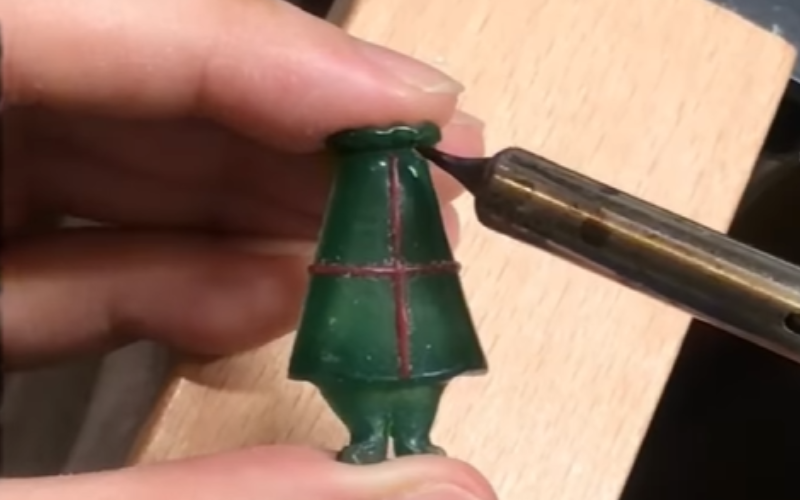 Close up of hands working at the bench. Left hand is holding a piece of green wax shaped like a torso and legs, right a wax heating pen is adding details