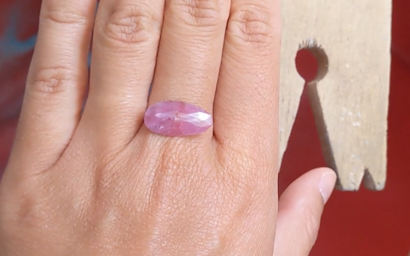 Close up of hands with a pink oval stone placed horizontally over the middle finger.