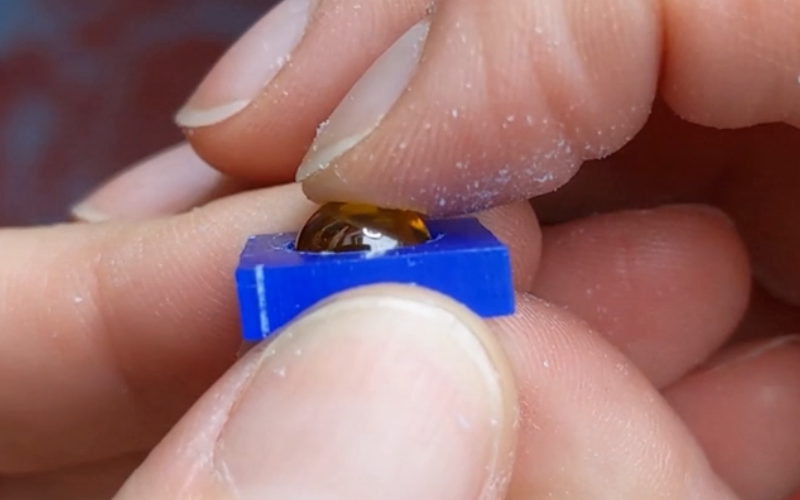 Close up of hands holding a piece of blue wax with a yellow round cabochon on top. The cabochon is fully inside the opening that's been carved in the wax