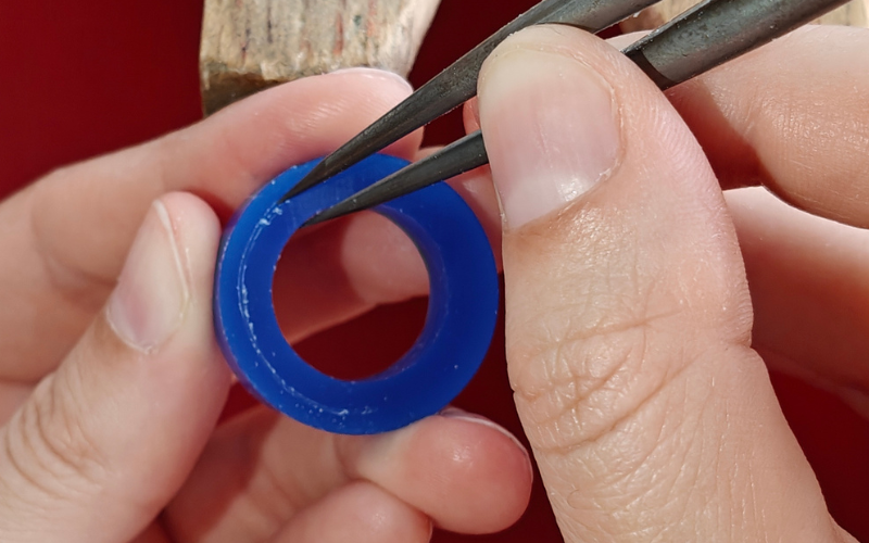 Close up of hands holding a piece of blue ring wax. Right hand is marking a line on the ring. The inside of the ring is used as the guide, already marked line is on the left