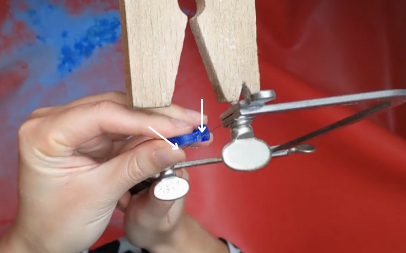 Close up of hands piercing on a blue wax ring. An arrow is pointed at the place where you're piercing and looking. Another arrow is pointing lower down where you might accidentally pierce something you don't want if you don't angle your saw enough