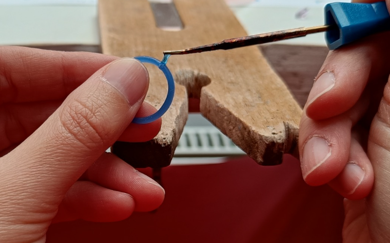 Close up of hands working at the bench. the left hand holds a blue wax ring and the right hand holds a wax heating pen and adds some extra blue wax on the ring