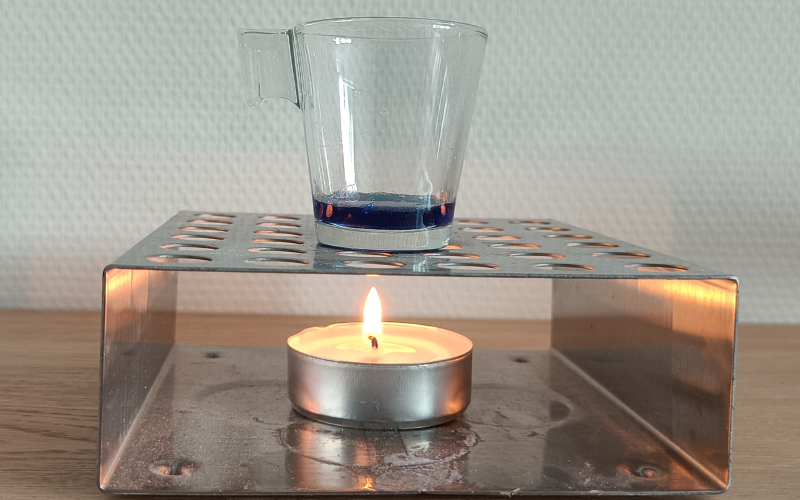 Burning tealight in a large metal holder. On top of tea light holder is a small glass with liquid blue wax inside