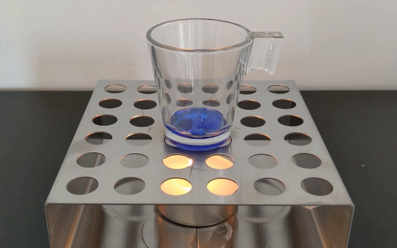  Close up of a small glass on top of a tealight. Blue wax is in the glass and it's half melted half solid