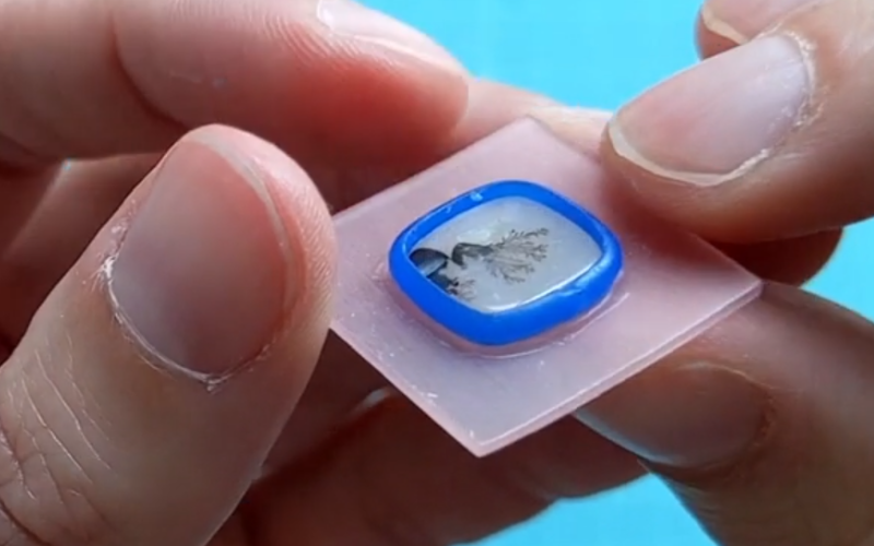 Close up of hands holding a setting from pink sheet wax and blue wire wax with a square dendrite on top. Around the blue wire you can see a clean line where wire and sheet are melted together
