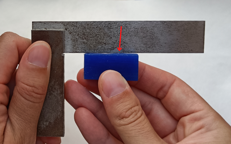 Close up of hands holding a set square and measuring a blue piece of wax against the thin arm. There are several gaps visible. A red arrow points to a very small gap that is not as easily visible