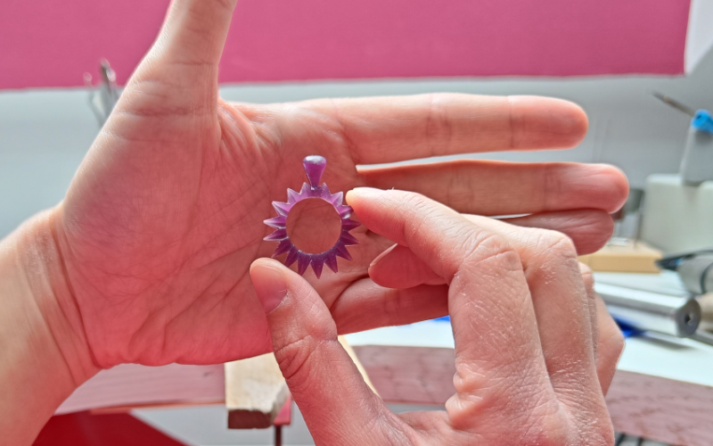 Close up of hands holding a pendant from purple wax. One hand is placed behind the piece to block immediate distractions from the bench making it easier to concentrate on the piece
