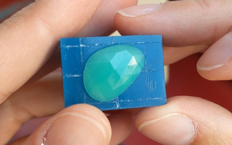 Close up of hands holding a piece of turquoise wax. Different centre lines are marked on the wax and a green stone is placed diagonally on it.