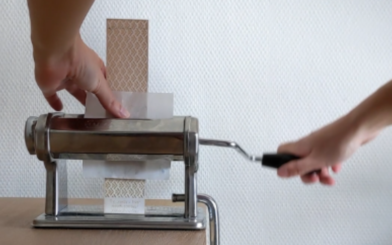 Close up of a pasta roller with a laser etched texture paper and a piece of sheet wax being rolled through