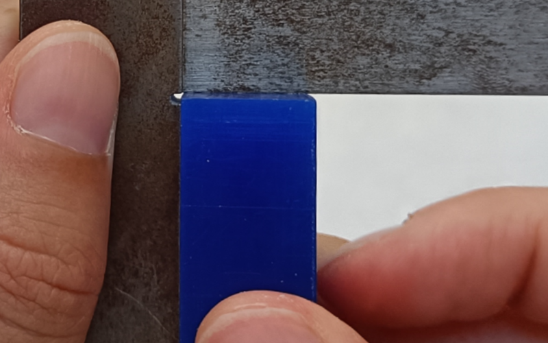 Close up of hands holding a set square and measuring a blue piece of wax in the corner. Both sides are touching 