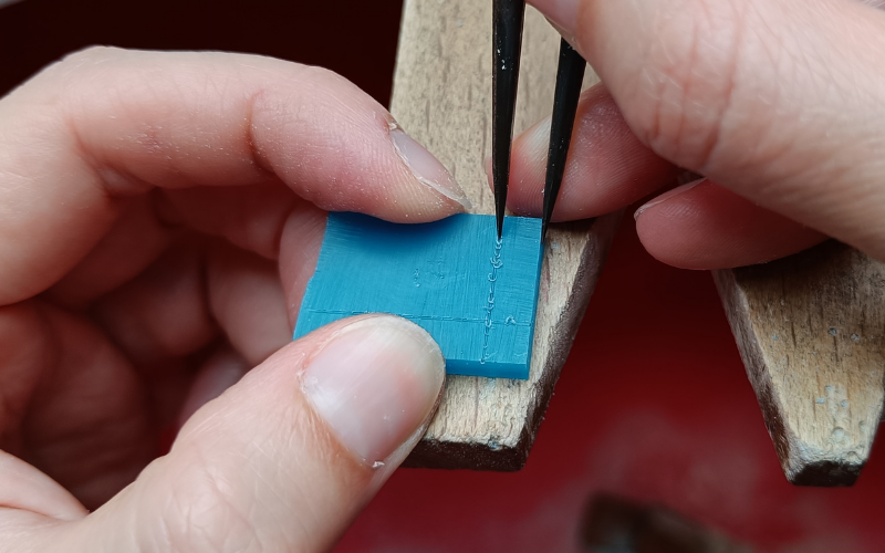 small rectangle of turquoise wax on a bench peg and hands marking a line with dividers on it 