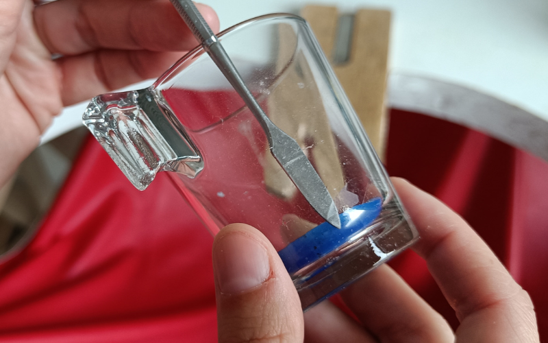 Close up of glass with blue wax in the bottom. A thin carving tool is pushed between glass and wax