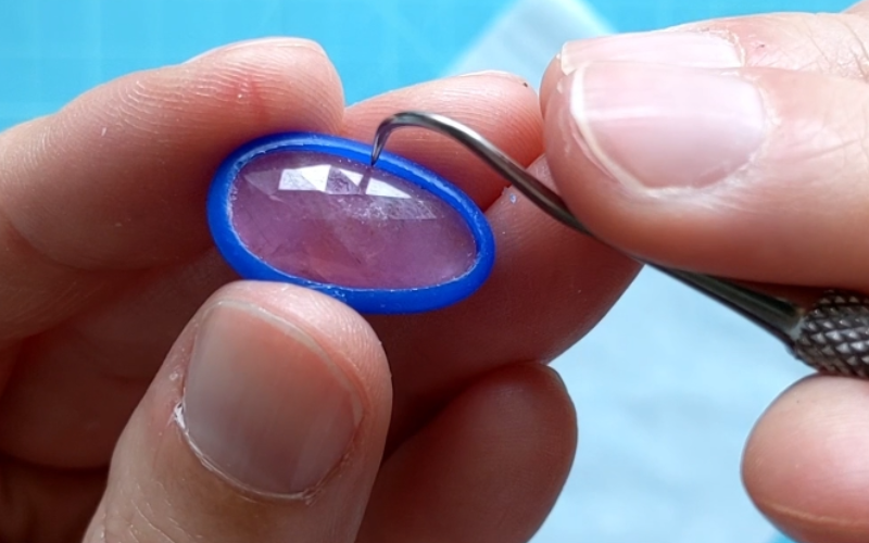 Close up of hands holding a pink stone with a blue wax wire bezel. A hook carving tool is gently pushing the walls outward