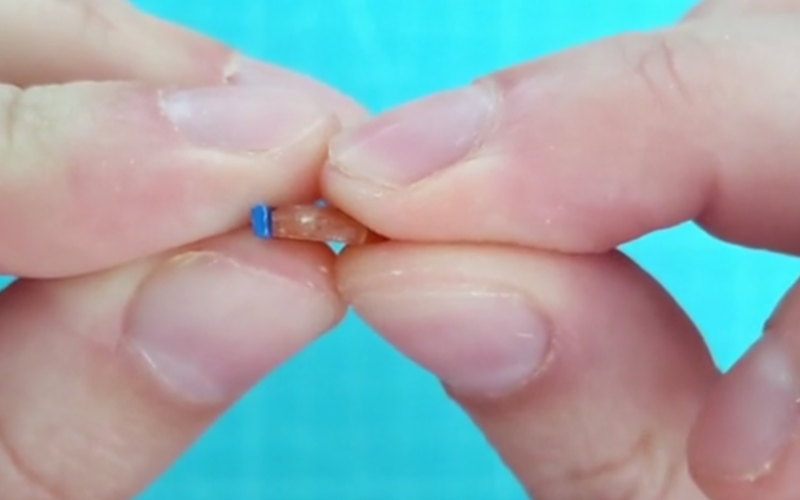 Close up of hands holding a sunstone showing the side profile with a piece of blue wax wire next to it showing of the flat rectangular shape of it
