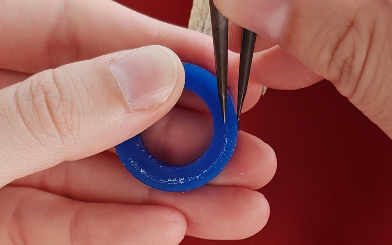 Close up of hands holding a piece of blue ring wax. Right hand is marking a line on the ring. The outside of the ring is used as the guide, already amrked line is on the right