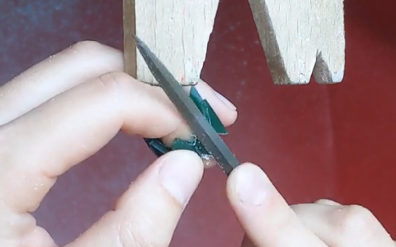 Close up of hands working at the bench. Left hand is holding a green wax ring, the ring finger is inside the ring to support the part that the right hand is filing. 