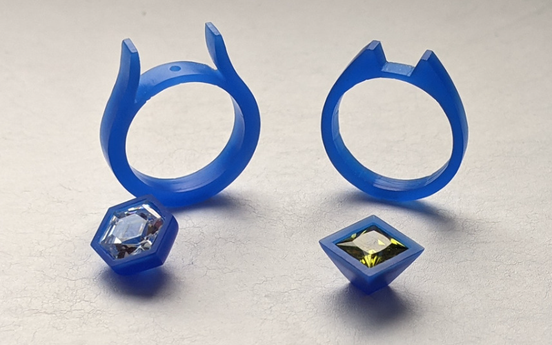 2 rings from blue wax with a little cut out in the centre for a setting to fit. In front of the left ring is a setting from blue wax with a hexagon shaped cz, in front of the right ring is a setting from blue wax with a square green cz 