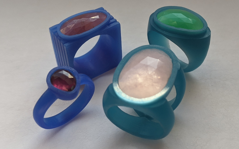 4 rings in wax with gemstones. 1 Small ring in blue wax with a red stone, one pink stone is a square ring from blue wax. One green stone in a rounded turquoise wax ring and one big pink stone in a turquoise wax ring with a split shank