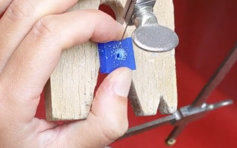Close up of hands working at the bench. The left hand is holding a piece of blue wax and the right hand is piercing the wax