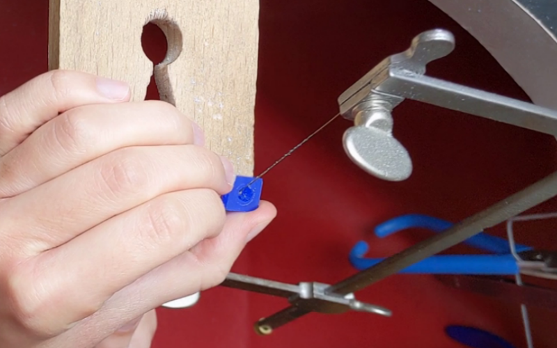 Left hand holding a piece of wax with a round cut out against a bench peg. Right hand holds a saw and is piercing the inside shape.