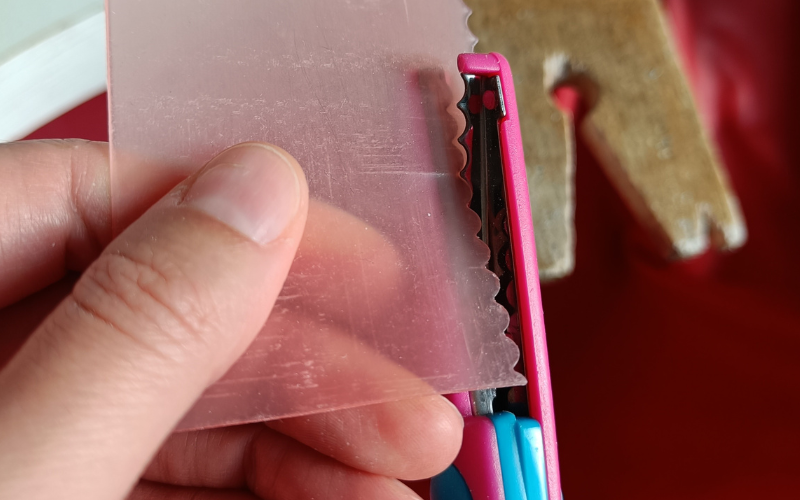 Close up of hands cutting a piece of pink sheet wax with scalloped edge scissors